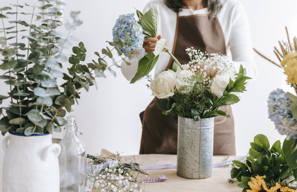 Crop unrecognizable female florist arranging hydrangea flowers into vase with roses while standing at table with assorted plants in room