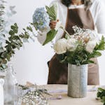 Crop unrecognizable female florist arranging hydrangea flowers into vase with roses while standing at table with assorted plants in room