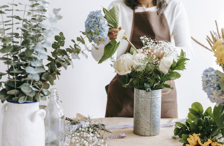 Crop unrecognizable female florist arranging hydrangea flowers into vase with roses while standing at table with assorted plants in room
