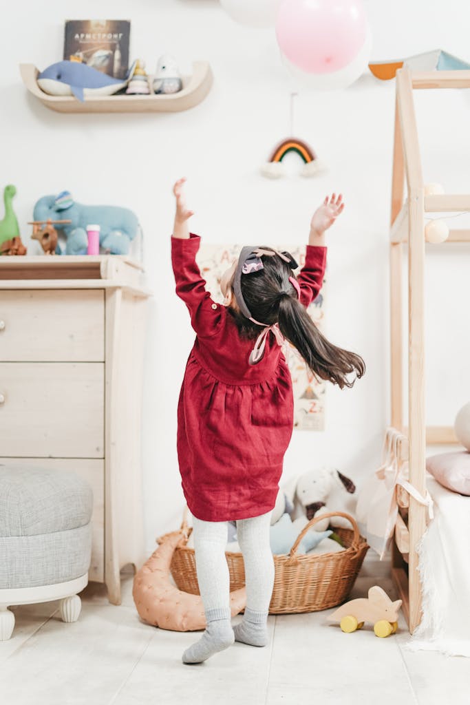 A young girl in a red dress jumps to catch balloons in a cozy bedroom setting.